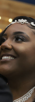 Bride in a white wedding gown holds onto groom’s arm as they stand together in an elegant indoor portrait with soft window light and dark interior background.