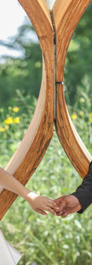 Bride wearing an ivory gown and holding a peach rose bouquet reaches for groom in a black suit under a wooden arch in an outdoor garden setting.