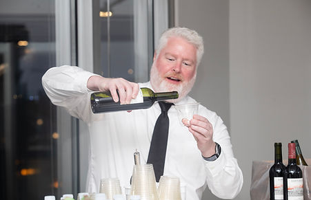 Man pouring wine behind a bar at an indoor event while preparing drinks for guests