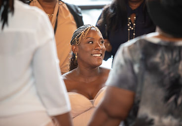 Expectant mother smiles during her maternity celebration, wearing a white strapless dress with braided hair, gold hair jewelry, and cross earrings as guests surround her.