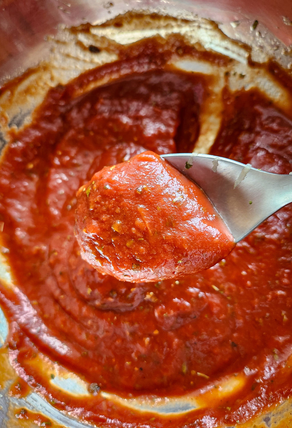 A small glass jar of bright red marinara sauce surrounded by fresh herbs and a clove of garlic on a wooden board.