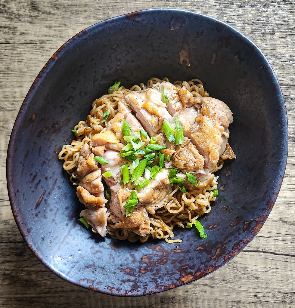 A bowl of glossy stir-fried tom yam instant noodles topped with sliced crispy skin-on chicken thigh, kaffir lime leaves, lemongrass, and chopped scallions.