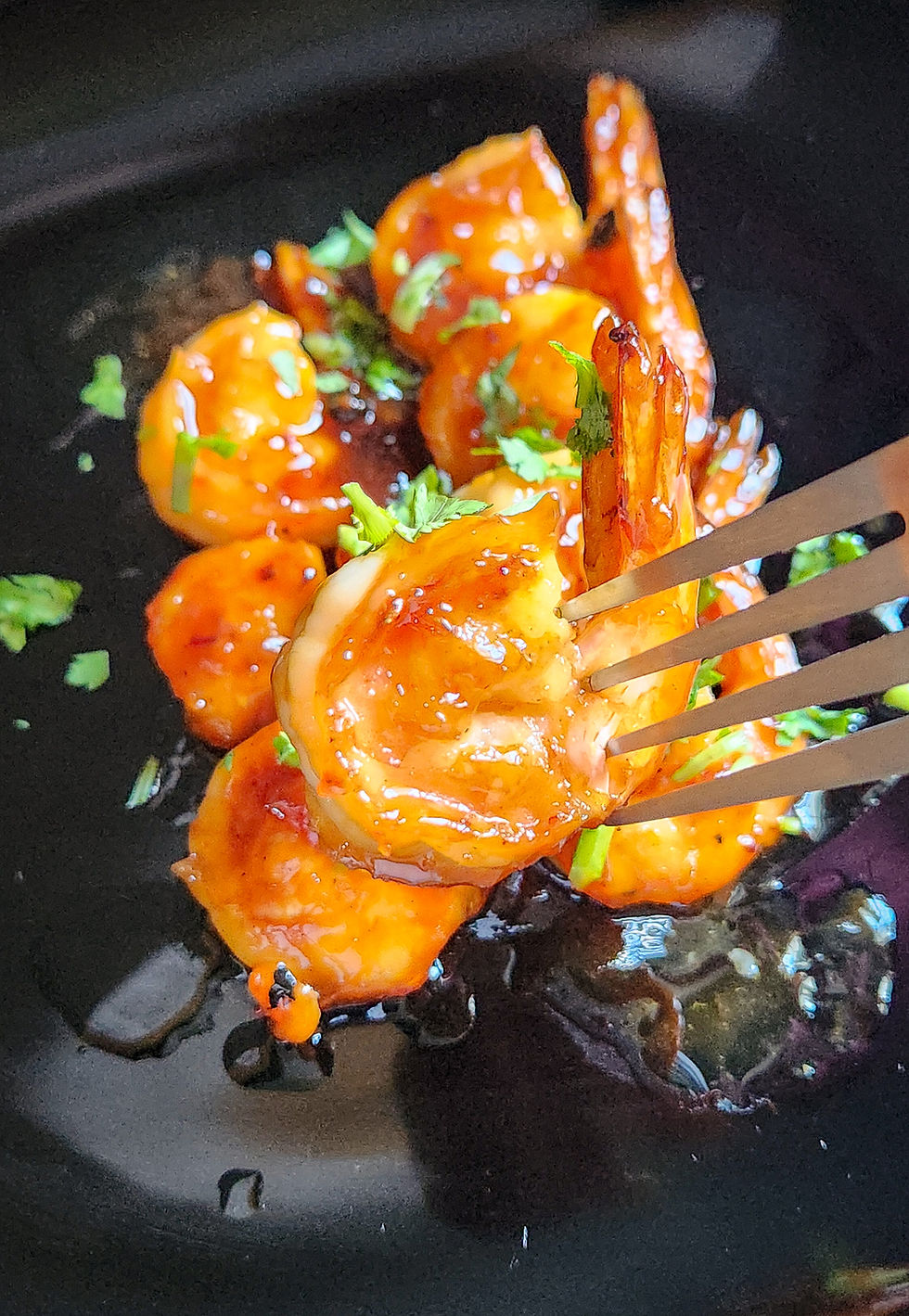 A fork lifting a single glazed prawn with coriander garnish over a black plate of prawns.