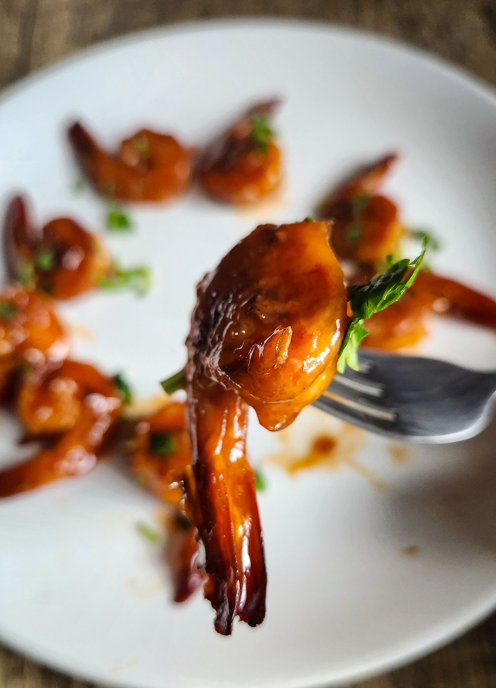 A single honey-glazed prawn on a fork held above a white plate with more prawns in background.