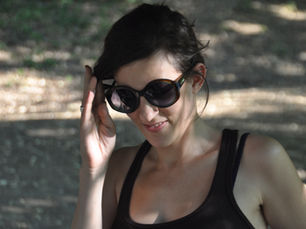 A woman outdoors wearing sunglasses, lightly touching the frame, with dappled light and trees in the background.