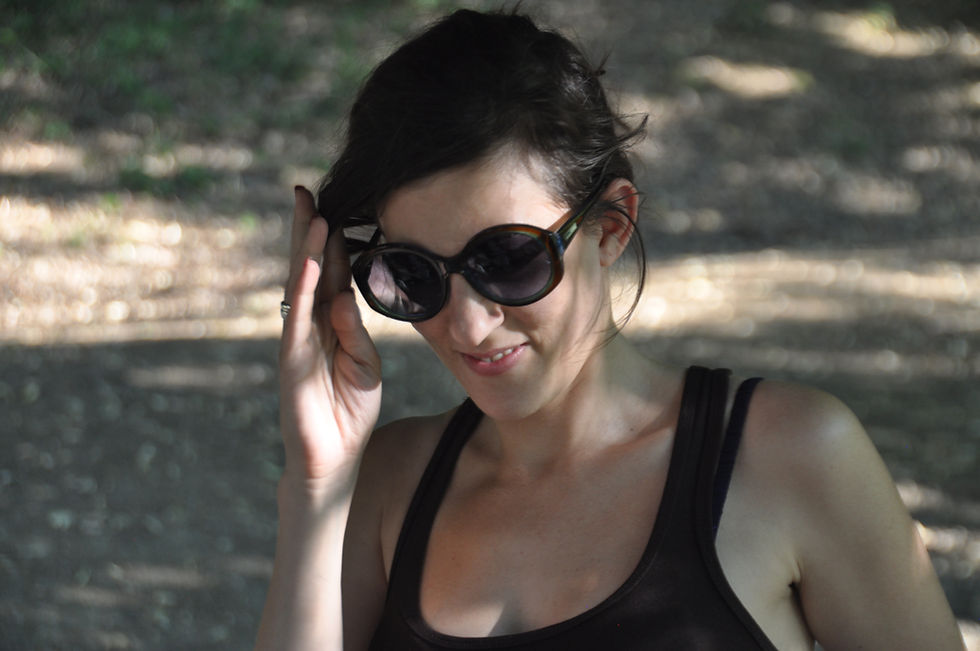 A woman outdoors wearing sunglasses, lightly touching the frame, with dappled light and trees in the background.
