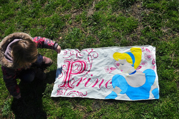 A child crouching on grass beside a hand-painted fabric featuring an illustrated character and decorative lettering.