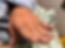 An elderly hand with long nails and gold rings rests on a light blue cloth. The background shows soft, patterned fabric.