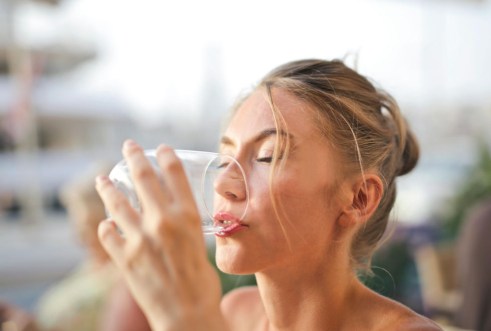 Woman drinking from clear glass with eyes closed, blurred outdoor background. Bright, relaxed mood.