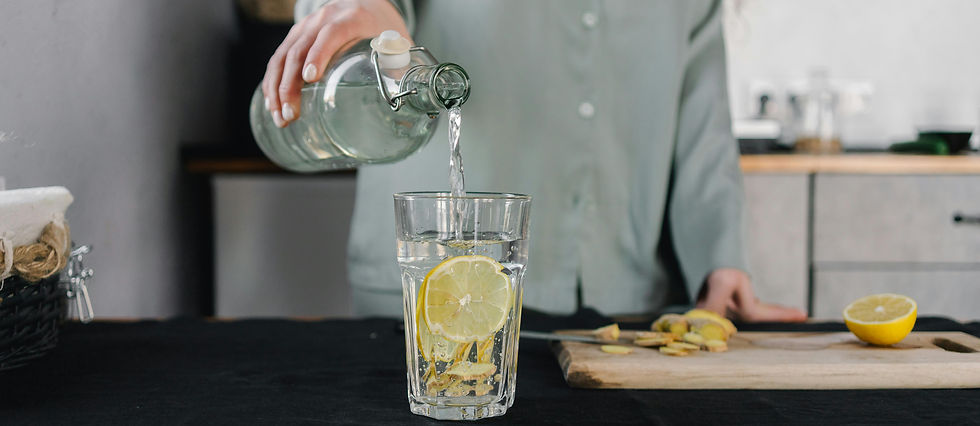 A person in a green shirt pours water from a bottle into a glass with lemon slices. A cutting board with ginger and lemon is in the background.