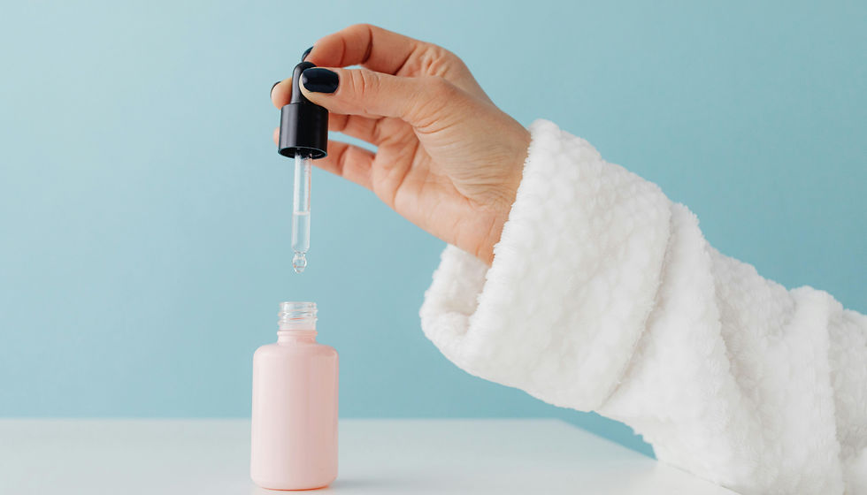 A hand in a white robe holds a dropper above a pink bottle against a blue background, with a drop of liquid forming at the tip.