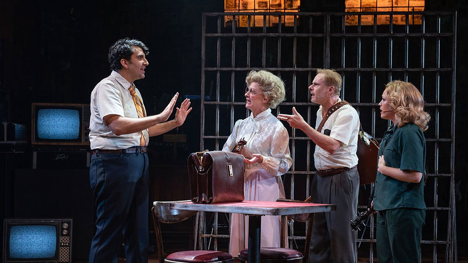 Four performers act around a table in a prison scene from Run Bambi Run at Milwaukee Repertory Theater, framed by tall jail bars and vintage television sets while focused theatrical lighting and zactrack SMART tracking keep the actors clearly illuminated.