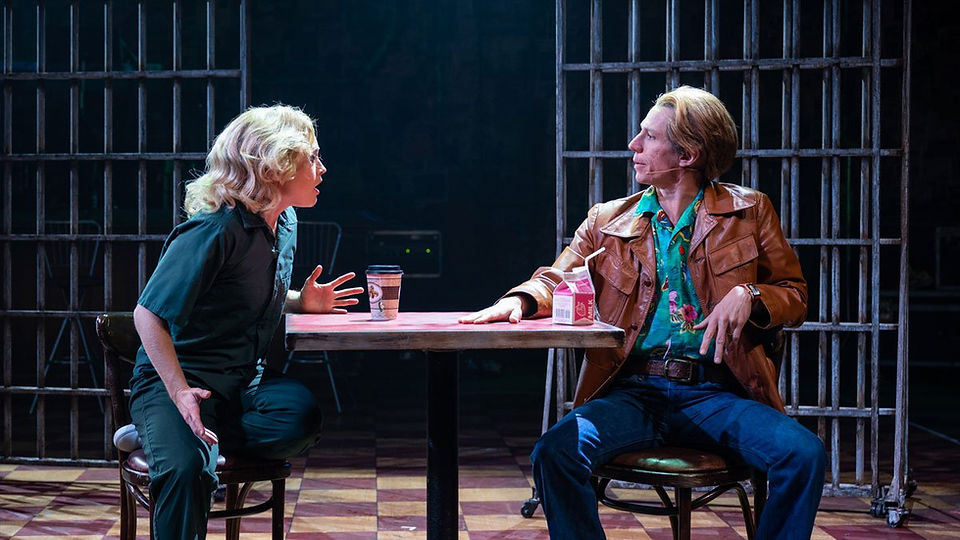 Two performers face each other across a small table during a prison scene in Run Bambi Run, surrounded by dark jail bars and cool focused lighting, with zactrack SMART supporting accurate automated tracking as the actors move through the theatrical space.