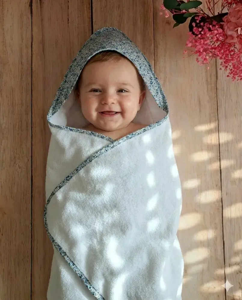 Bébé souriant enveloppé dans une cape de bain artisanale en éponge blanche avec capuche en coton Liberty, création fait main pour un trousseau de naissance doux et raffiné.