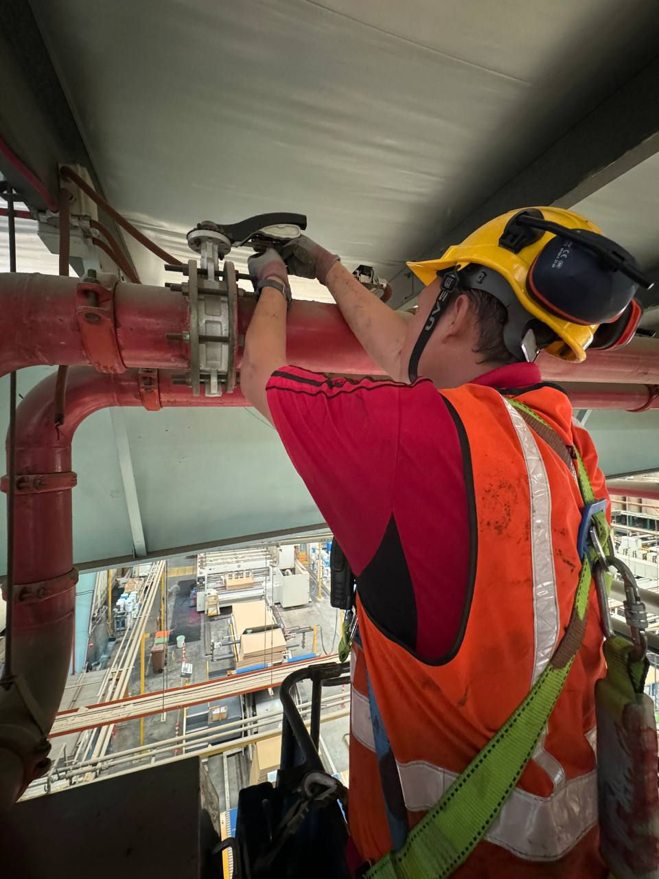 A fire contractor, equipped with a hard hat and safety harness, conducts maintenance on a fire suppression system pipe, high above the floor of an industrial facility.