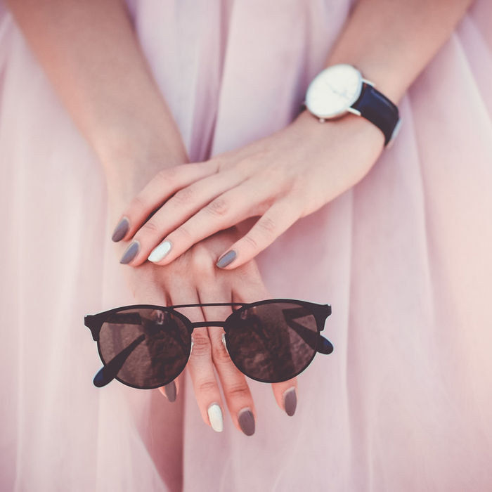 Woman's hands holding sunglasses, wearing watch, pink dress, and soft background.