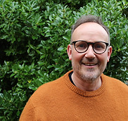 Man - Paul Heseltine - smiles in front of green bushes wearing glasses and orange sweater.