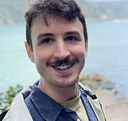 Smiling man - Ilias Bloomfield - with a moustache in front of a scenic ocean background.