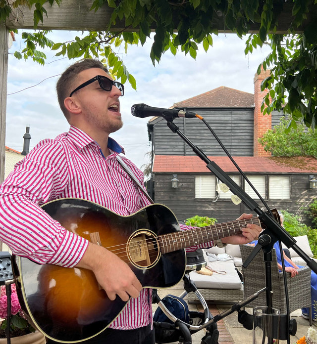 Tom Ryder singing outdoors under a pergola in a pink striped shirt