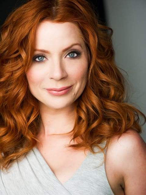 Smiling red-haired woman in a light gray top, posed against a plain gray background.