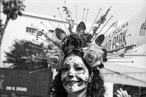 Woman in elaborate Day of the Dead makeup with floral headpiece and sign.