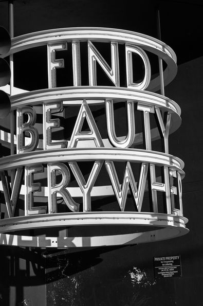 Neon sign says: FIND BEAUTY EVERYWHERE. Roads Less Traveled, in city streets.