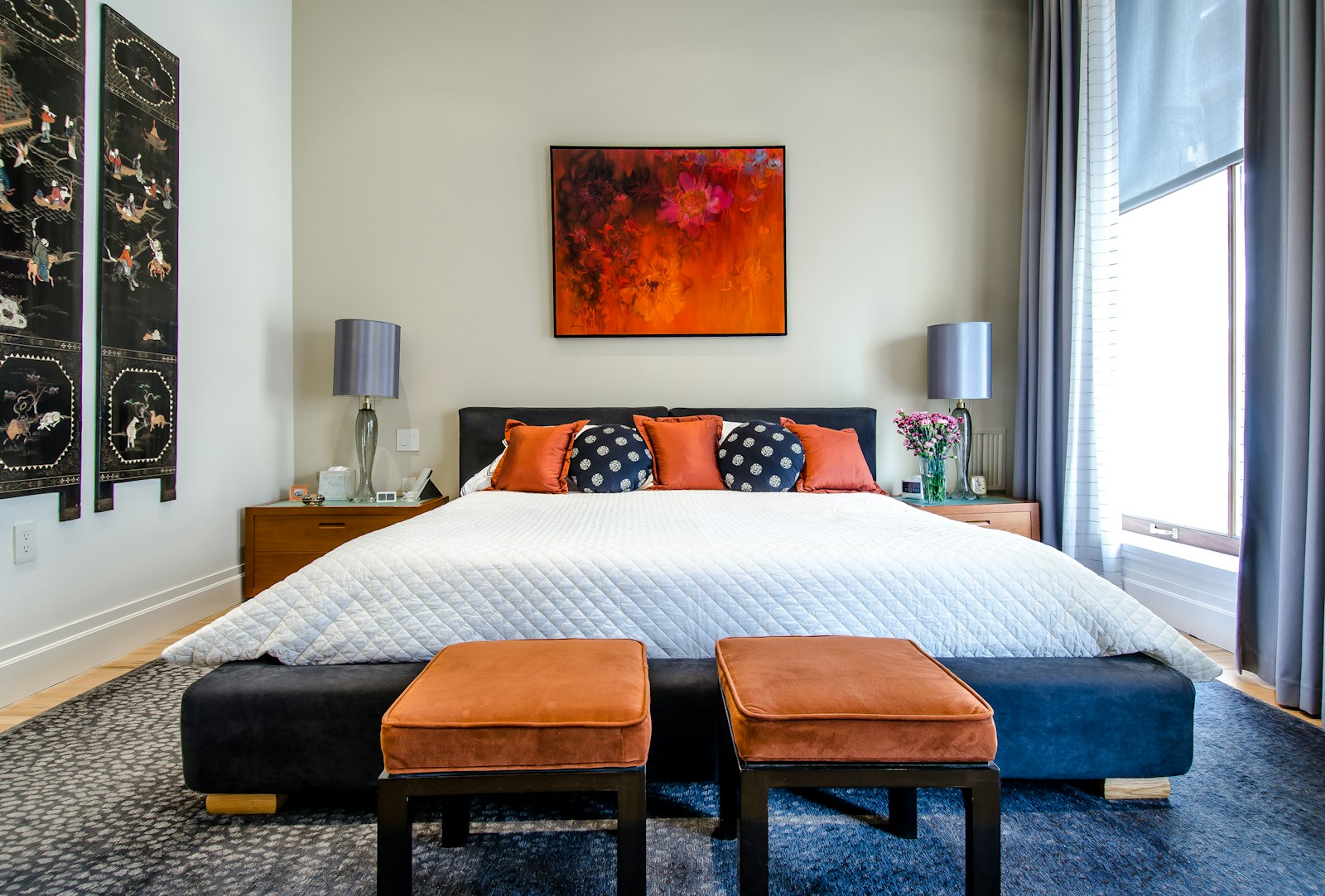 Bedroom with orange pillows, two benches, and a floral painting, no people