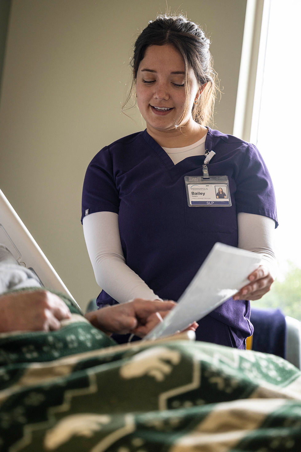 A nurse in purple scrubs assists a person in a hospital bed, holding a document. The room is softly lit, and both appear calm.