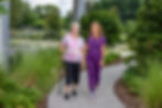 Elderly woman with a cane walks beside a smiling nurse in purple scrubs on a garden path, surrounded by lush greenery and a railing.
