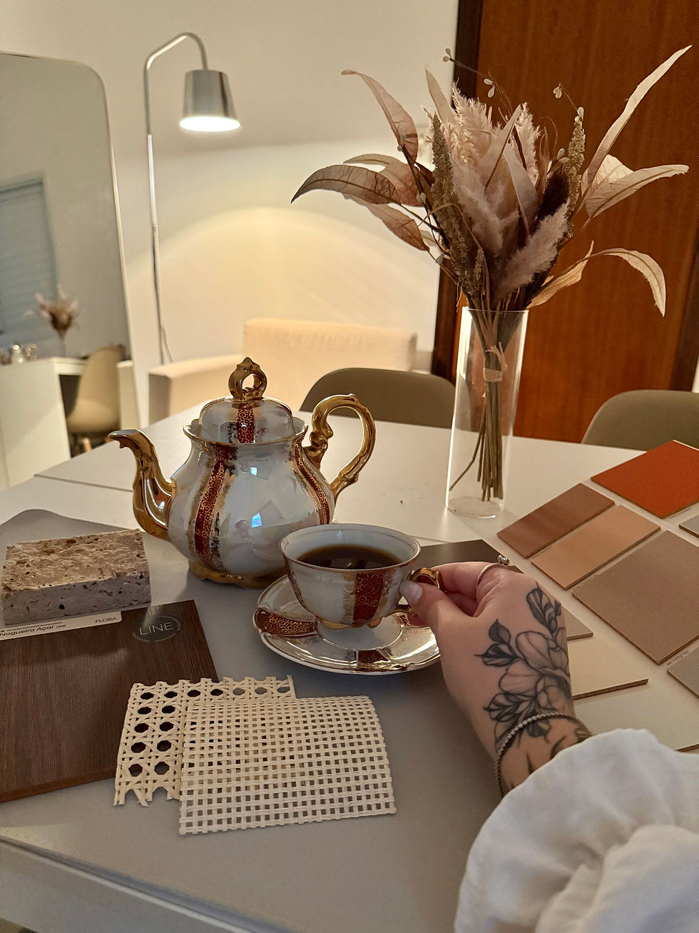 A tattooed hand holds a tea cup by a teapot on a table with fabric swatches. Warm lighting, dried flowers in a vase, and cozy ambiance.