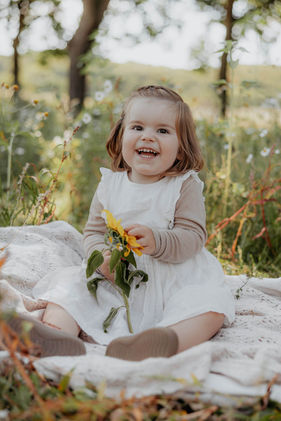 Lille pige sidder i græsset og holder en solsikke under naturlig børnefotografering i naturen nær Aarhus.