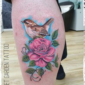 Tätowierter kleiner Vogel, sitzend auf einer pinken Blüte mit Schnörkel.