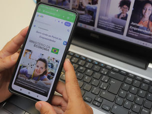 Uma mão segurando um smartphone que exibe uma página web com informações sobre empresas e profissionais. A pessoa está usando um teclado de computador ao fundo, sugerindo que ela pode estar trabalhando em um ambiente de escritório ou de trabalho remoto.