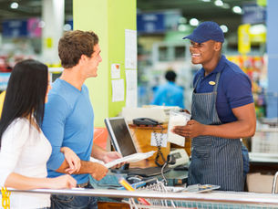 Na imagem, há uma loja com três pessoas. Um homem está atrás do banco de vendas, vestindo um uniforme azul e segurando um bilhete. Ele está sorrindo para a câmera. Dois outros indivíduos estão em frente ao banco de vendas: um deles está segurando uma caixa de dinheiro e o outro está segurando uma nota ou documento. Todos parecem estar em uma loja com produtos visíveis no fundo.