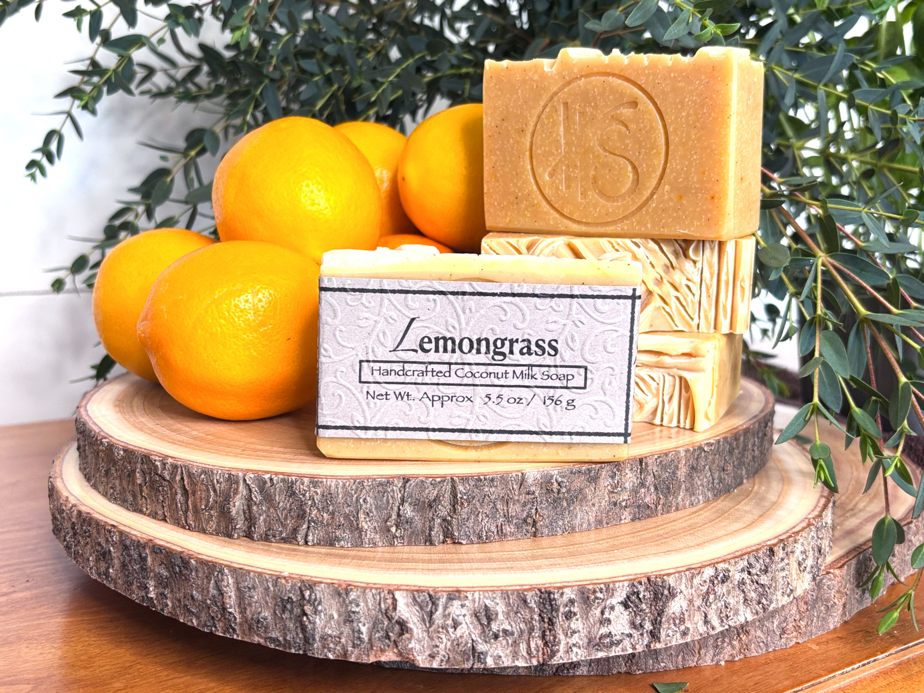 Four yellow bars of Lemongrass soap with embossed label on top of stacked wooden slabs surrounded by lemons and green leaves.