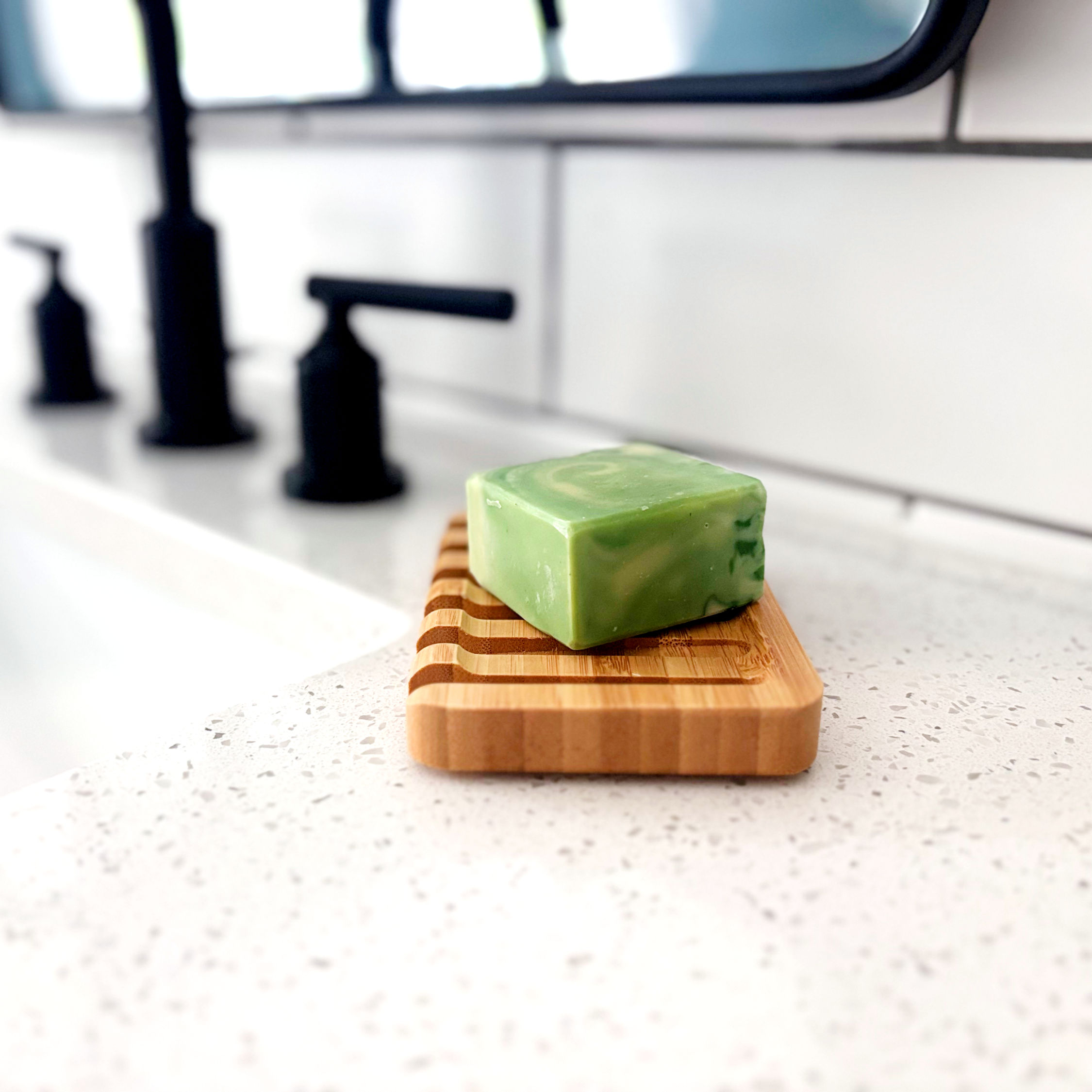 Green & cream colored bar of soap sitting on a wooden waterfall soap dish next to a sink.