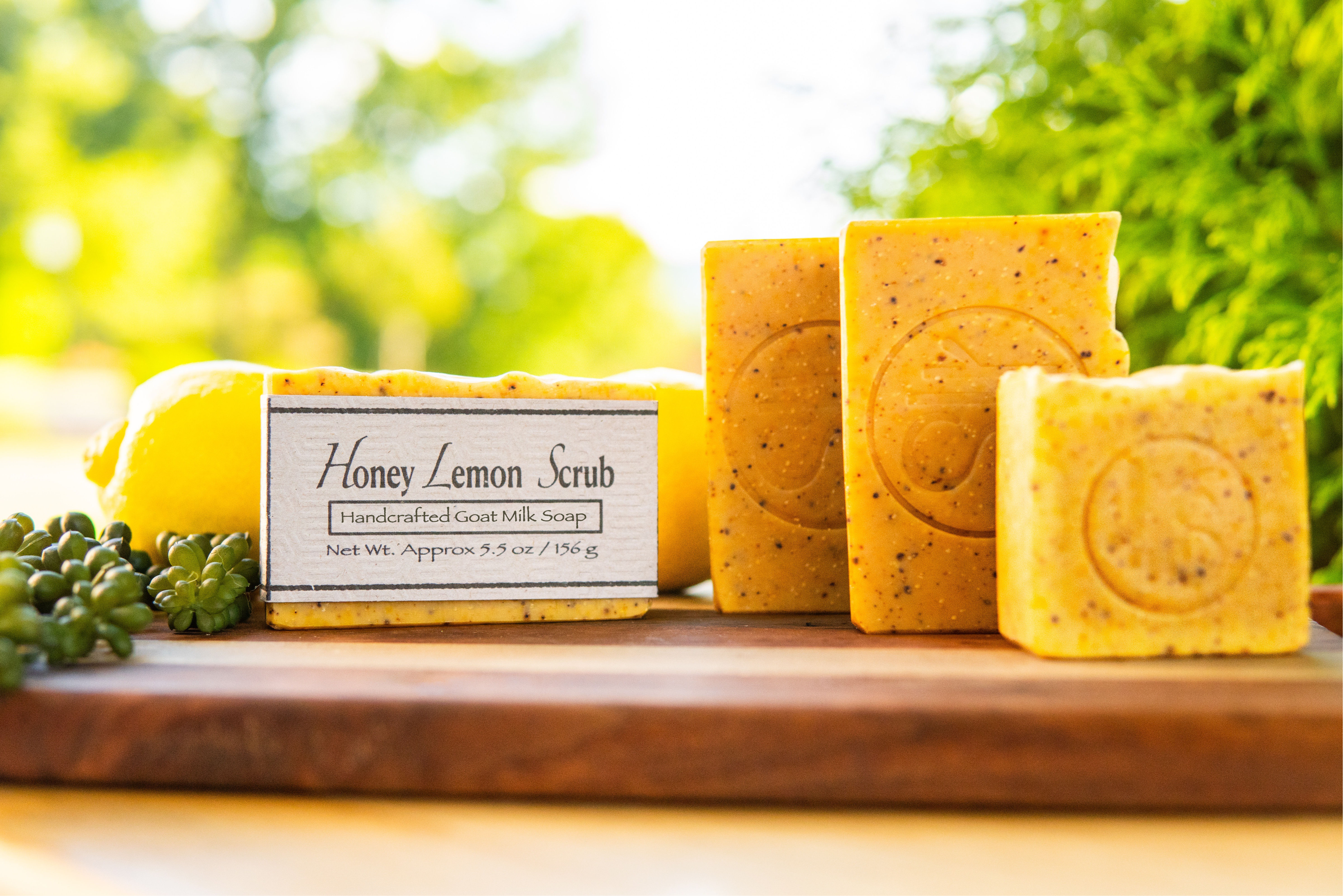 Four yellow bars of Honey Lemon Scrub soap with embossed label sitting on wood board with lemons and honey.