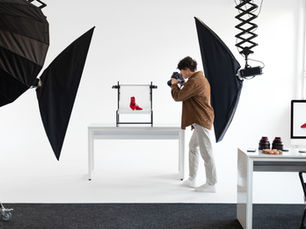 A photographer is taking a photo of a pair of red high-heeled shoes in a professional studio setup. The shoes are placed on a white background between two large softbox lights. A computer monitor on the right displays the captured image of the shoes. Various camera lenses and a plate of macarons are on the nearby desk.