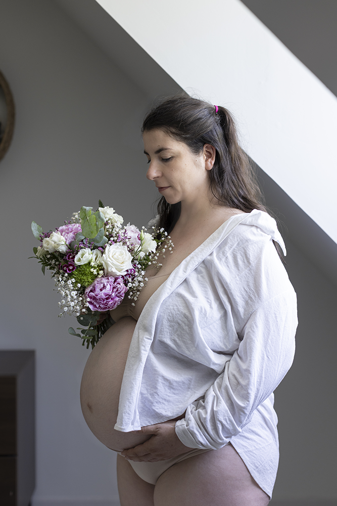 La future maman est vêtue d'une chemine et tient un bouquet de fleurs chez elle lors de sa séance grossesse