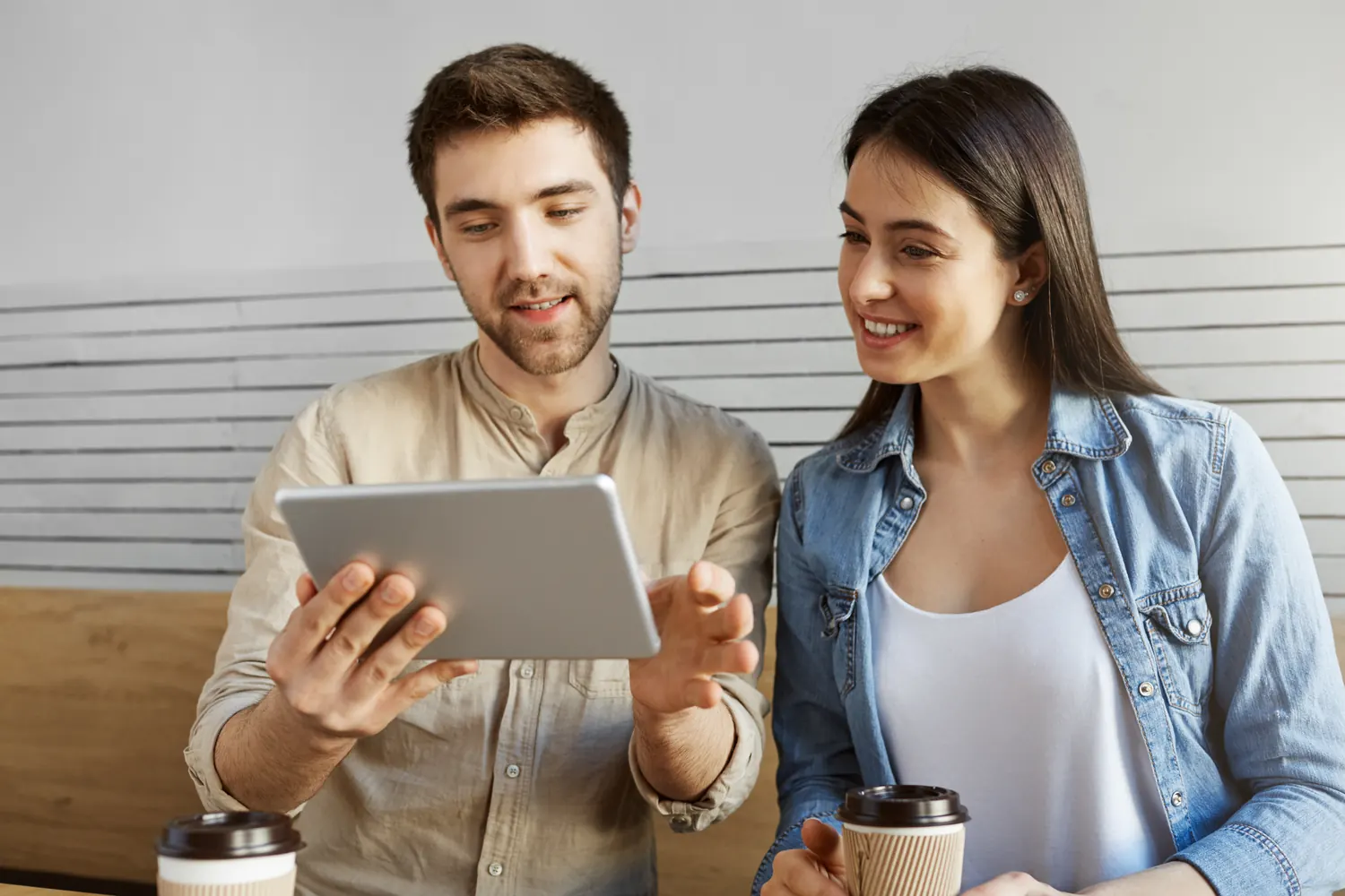Mann und Frau sitzen mit Kaffeebechern nebeneinander und sehen gemeinsam auf ein Tablet
