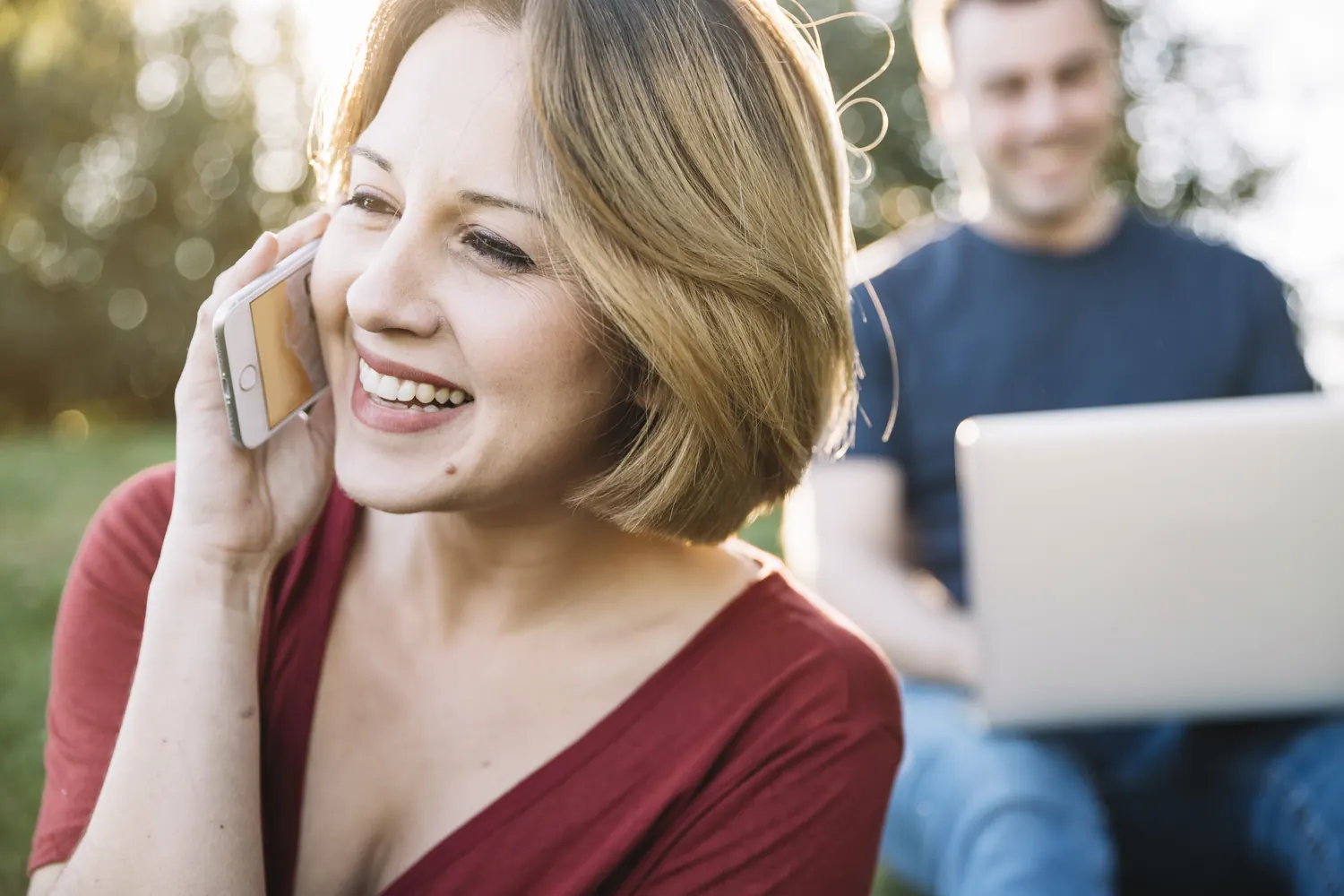 Lächelnde Frau telefoniert im Freien mit dem Smartphone, im Hintergrund ein Mann am Laptop