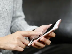 Manos usando un smartphone rosa sobre fondo oscuro; persona con suéter gris tocando la pantalla.