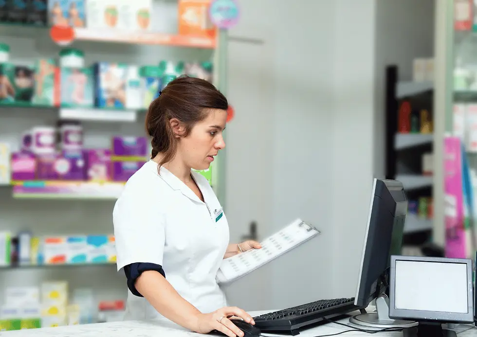 Farmacéutica con bata blanca revisa una lista en portapapeles junto al ordenador en una farmacia, con estantes de medicinas de fondo.