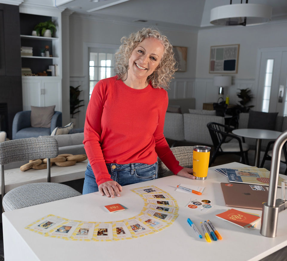 Woman with curly grey hair and subtle makeup standing at her kitchen counter with tarot cards