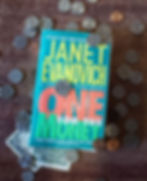 The book One for the Money by Janet Evanovich is laying on a wooden background surrounded by coins. A one dollar bill is tucked under the book's lower left corner.