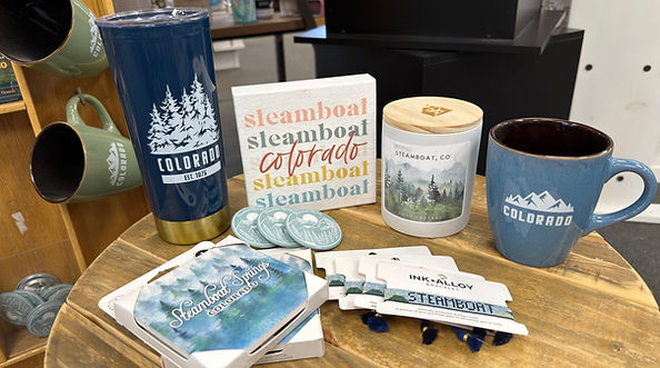 Teal Colorado tumbler and blue mug, Steamboat Springs plaque and coasters, candle, bracelet and magnet display showcasing available souvenirs.