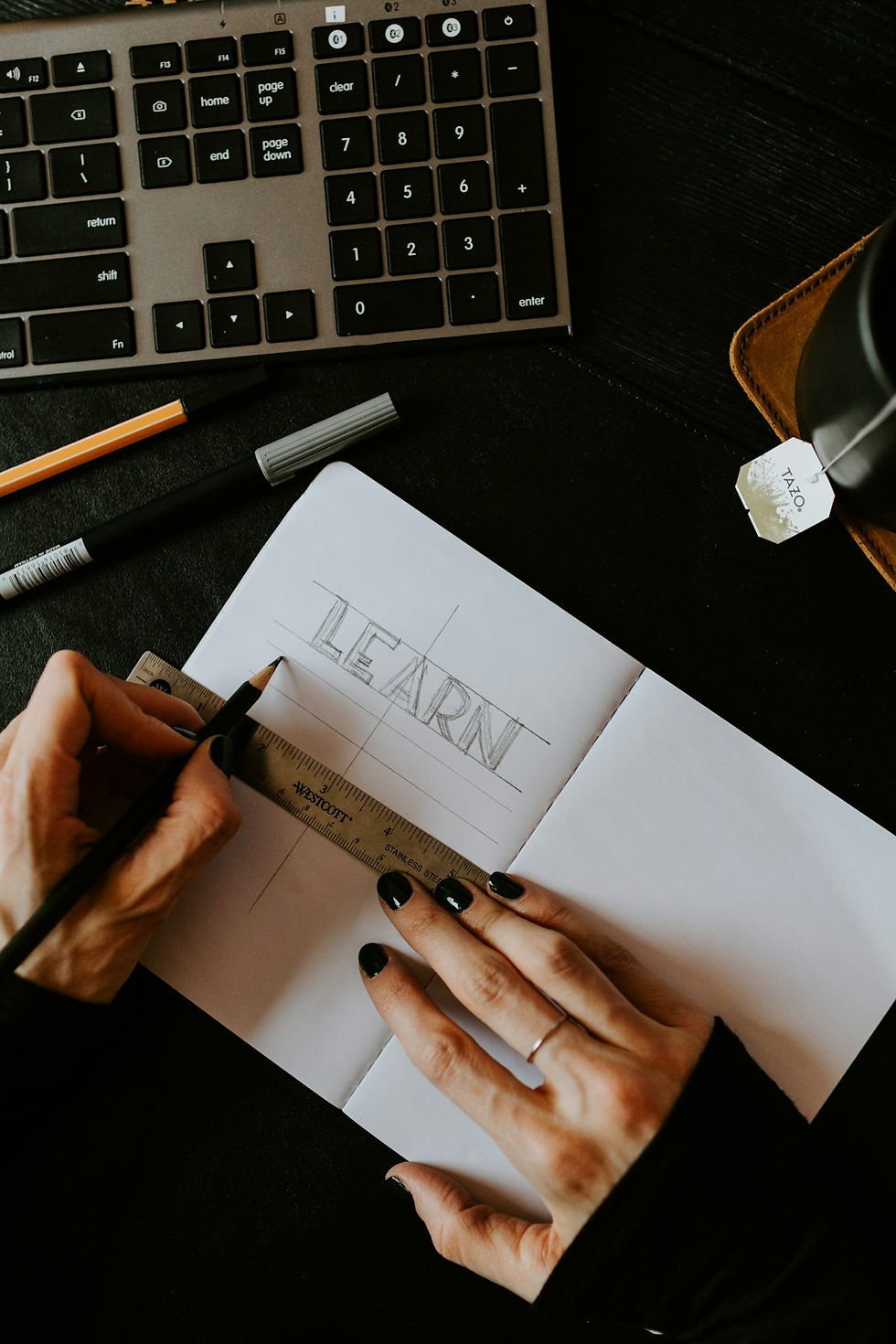 a laptop keyboard and pencils and pens and a notebook with two hands and a ruler writing LEARN