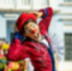 A clown in full makeup and costume with hands on their head looking at the camera
