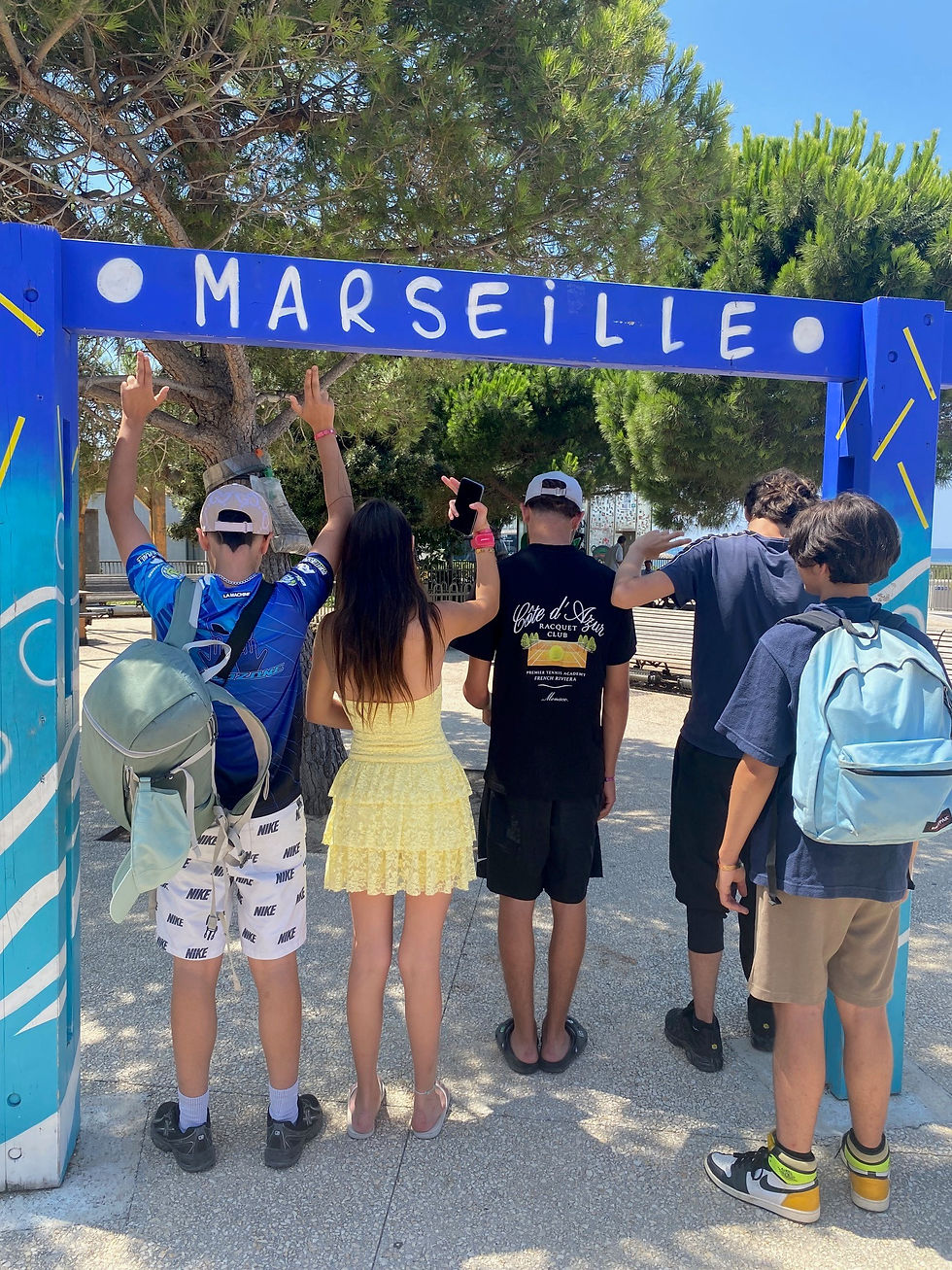 Cinq ados de dos sous une arche bleue MARSEILLE, bras levés, dans un parc ensoleillé bordé de pins.