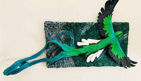 3D sculpture of a quetzal bird flying over a rectangle textured canvas of green foliage, river, and forest fires.  Quetzal's wings and tail outstretch majestically past the edges of the canvas.
